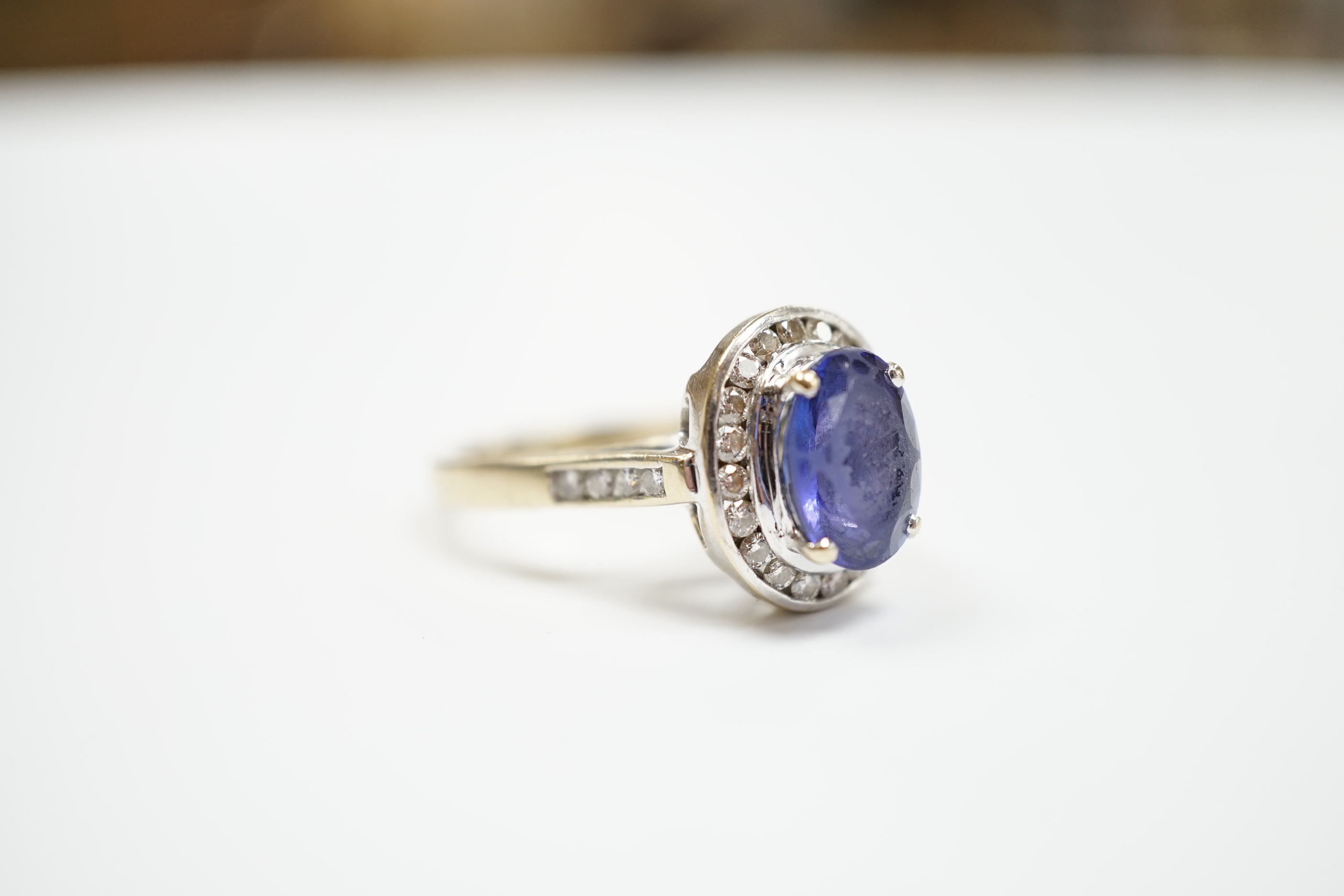 A modern 750 and oval cut single stone tanzanite(worn) set dress ring, with diamond set border and shoulders, size P, gross weight 8.9 grams.